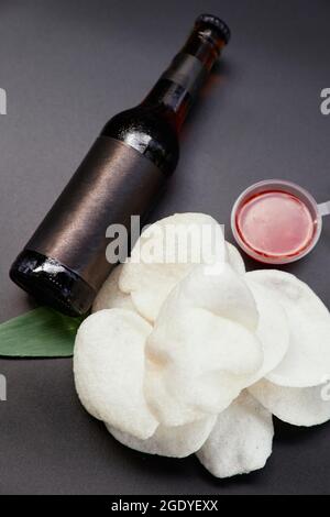 Reischips mit süßer und saurer oder würziger Chilisauce mit dunkler Flasche frischem Getränk oder Bier. Junk-Food- oder Fastfood-Konzept. Draufsicht auf Krupuk-Chips auf schwarzem Hintergrund. Hochwertige vertikale Fotos Stockfoto