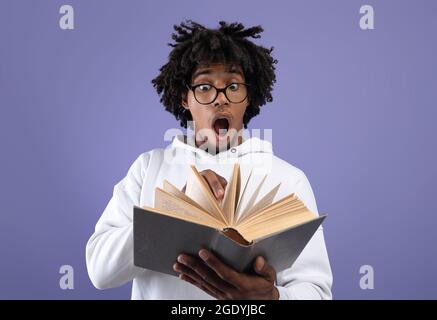 Schwarzer Teenager-Student, der Lehrbuch unter Schock ansieht, Angst vor zu viel Hausaufgaben auf violettem Studiohintergrund Stockfoto