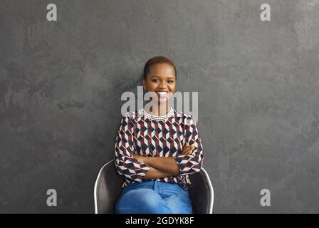 Porträt einer attraktiven jungen dunkelhäutigen Frau, die auf einem Stuhl sitzt und aufrichtig lächelt. Stockfoto