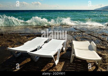 Eine Reihe von drei Sonnenliegen mit Blick auf das Meer. Sonniger Strand mit drei Sonnenliegen. Das Meer bei stürmischem Wetter. Wellen stürzen auf die Brüstung. Stockfoto