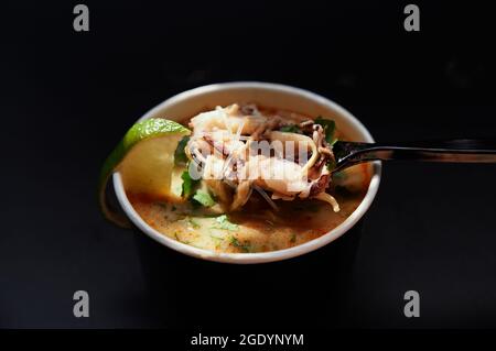 Löffel und Schüssel mit Nudelsuppe mit Meeresfrüchten und Tintenfisch. Thailändische oder vietnamesische Küche: Tom Yum Goong würzige und saure Suppe mit leckeren Reisnudeln mit Oktopus auf schwarzem Hintergrund. Hochwertige Bilder Stockfoto