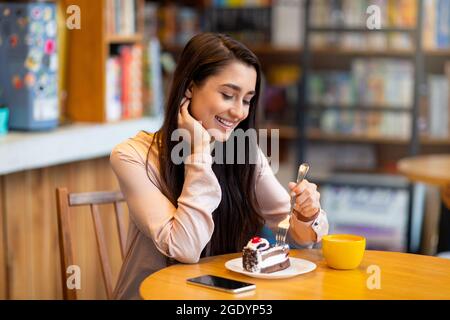 Positive Dame, die Kuchen und Kaffee trinkt, während sie im Café sitzt, Zeit in der Cafeteria verbringt, Platz für Kopien Stockfoto