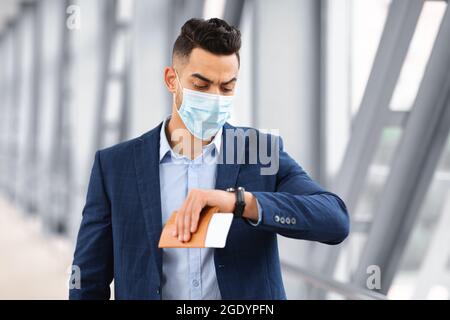 Arabischer Geschäftsmann in medizinischer Maske, der am Flughafen läuft und auf die Armbanduhr schaut Stockfoto