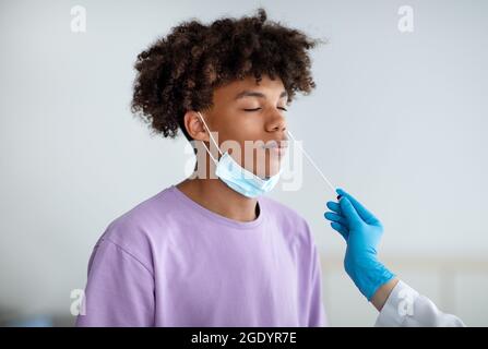 Arzt in Gummihandschuhen, die nasalen Coronavirus-PCR-Test für afroamerikanische Teenager, mit sterilen Abstrichstäbchen in Innenräumen Stockfoto