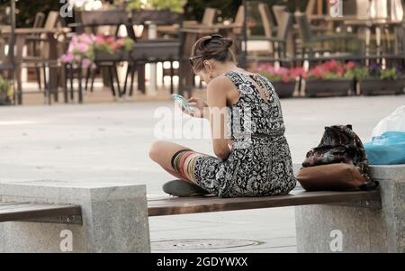 Junge Frau, die in Sofia, Bulgarien, auf der Bank sitzt und ihr Mobiltelefon anschaut Stockfoto