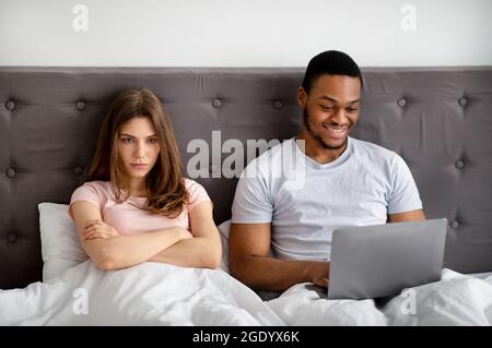 Der junge schwarze Mann, der einen Laptop im Bett benutzt, seine beleidigte Freundin, die zu Hause unter Vernachlässigung litt Stockfoto