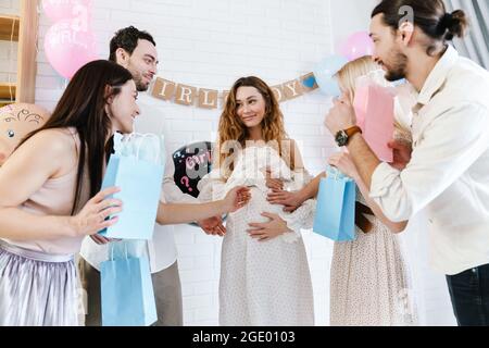 Junge, glückliche, schwanger Frau, die Geschenke von ihren Freunden während der Geschlechtsenthüllung in Innenräumen annimmt Stockfoto