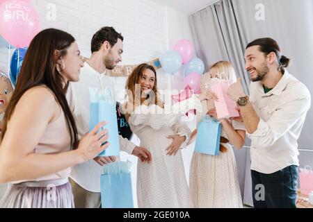 Junge, glückliche, schwanger Frau, die Geschenke von ihren Freunden während der Geschlechtsenthüllung in Innenräumen annimmt Stockfoto