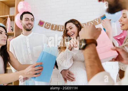 Junge, glückliche, schwanger Frau, die Geschenke von ihren Freunden während der Geschlechtsenthüllung in Innenräumen annimmt Stockfoto