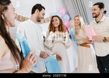 Junge, glückliche, schwanger Frau, die Geschenke von ihren Freunden während der Geschlechtsenthüllung in Innenräumen annimmt Stockfoto