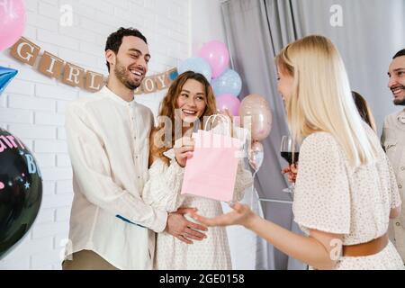 Junges glückliches Paar, das Geschenke von ihrer Freundin während der Geschlechtsenthüllung in Innenräumen annimmt Stockfoto