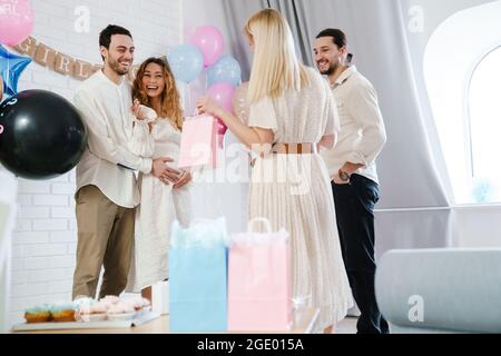 Junges glückliches Paar, das Geschenke von ihrer Freundin während der Geschlechtsenthüllung in Innenräumen annimmt Stockfoto