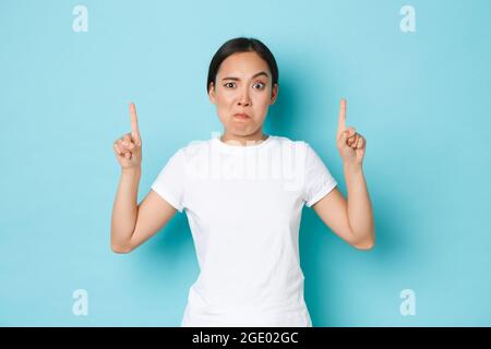 Wütend und unzufrieden, asiatische lustige Mädchen in weißem T-Shirt, so dass komplizierte, verdächtige oder verrückte Grimasse, warten Erklärung, zeigt die Finger nach oben auf Stockfoto