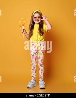 Niedliches verspieltes rothaariges Mädchen in Mädchenkleidung, gelbes T-Shirt, Leggings und Sneakers mit großen Lutscher-Süßigkeiten Stockfoto