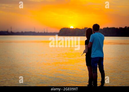 Silhouette eines Paares, das in der Abenddämmerung den Sonnenuntergang über dem Fluss genießt. Liebeskonzept. Stockfoto