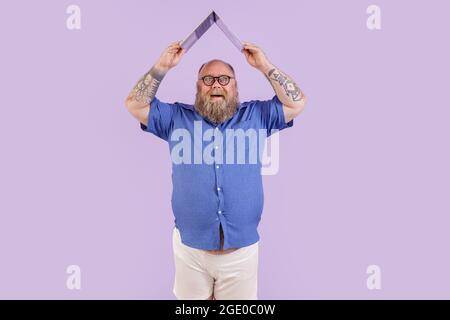 Fröhlicher Mann mit Übergewicht hält Laptop über dem Kopf als Dach auf violettem Hintergrund Stockfoto