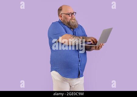 Ein emotionaler Mann mit Übergewicht arbeitet auf einem Laptop, der im Studio auf violettem Hintergrund steht Stockfoto