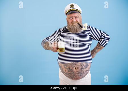 Schneidig fetter Seemann mit Bart und Rauchpfeife hält schaumiges Bier auf hellblauem Hintergrund Stockfoto