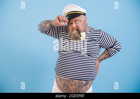 Reifer fetter Seemann mit Bart und Vintage-Pfeife passt Hut auf hellblauem Hintergrund an Stockfoto