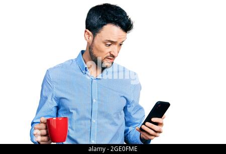 Hispanic Mann mit Bart mit Smartphone und trinken eine Tasse Kaffee ahnungslos und verwirrt Ausdruck. Zweifel Konzept. Stockfoto