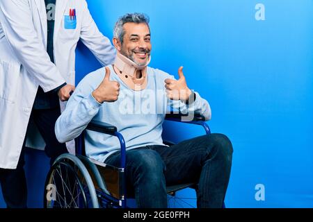Schöner Mann mittleren Alters mit grauen Haaren auf dem Rollstuhl trägt Halsband Erfolgszeichen tut positive Geste mit der Hand, Daumen hoch lächelnd und happ Stockfoto