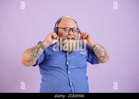 Glücklicher bärtiger Mann mit Übergewicht hört Musik mit Kopfhörern auf purpurem Hintergrund Stockfoto