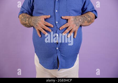 Mann mit Übergewicht und Tattoos hält die Hände auf einem großen Bauch auf violettem Hintergrund Stockfoto