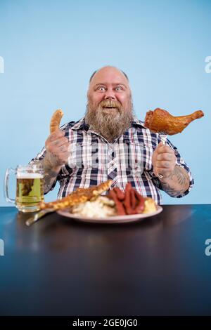 Fröhliche Person mit Übergewicht sitzt am Tisch mit fetten Speisen und Bier Stockfoto