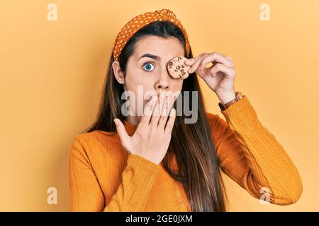 Junge Brünette Teenager hält Cookie über Auge bedeckt Mund mit der Hand, schockiert und Angst vor Fehler. Überrascht Ausdruck Stockfoto