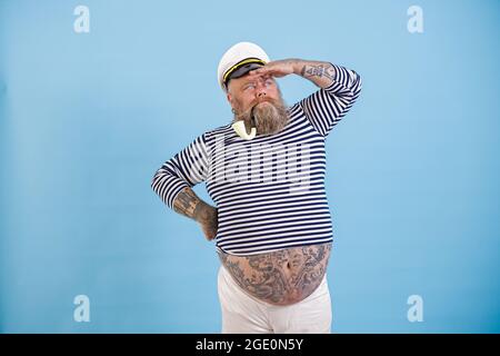 Plus Size Seemann mit Rauchrohr und Hut schaut in die Ferne auf hellblauem Hintergrund Stockfoto