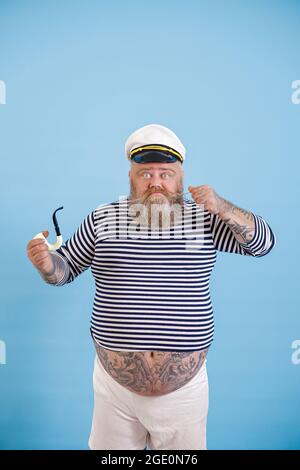 Bärtig übergewichtiger Mann Seemann mit Rauchrohr strafft Schnurrbart auf hellblauem Hintergrund Stockfoto