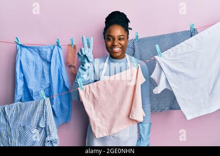 afroamerikanische Frau mit geflochtenen Haaren, die Kleidung an der Wäscheleine wäscht, lächelt mit einem glücklichen Gesicht, das die Kamera anwinkelt und mit den Fingern ein Siegeszeichen tut. Stockfoto