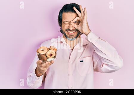 Kaukasischer Mann mittleren Alters, der süßes Gebäck hält, lächelt und lächelt und ein Schild mit der Hand auf dem Auge durch die Finger schaut Stockfoto