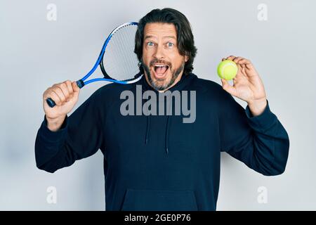 Kaukasischer Mann mittleren Alters spielt Tennis mit Schläger und Ball und feiert verrückt und erstaunt über den Erfolg mit offenen Augen schreiend aufgeregt. Stockfoto