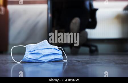 Eine medizinische Maske, die auf dem Boden eines Büros abgelegt wurde, mit den Füßen eines arbeitenden Mannes im Hintergrund. Nach einer Pandemie wieder an die Arbeit. Stockfoto