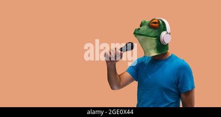Person in Froschmaske mit einem Mikrofon in der Hand und kabellosen Kopfhörern singen und genießen Karaoke auf orangefarbenem Hintergrund mit Kopierraum Stockfoto