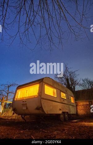 Alter weißer Wohnwagen-Anhänger mit gelber Beleuchtung im Hinterhof bei stary Blue Night Stockfoto