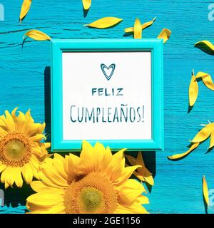 Text Feliz Cumpleanos bedeutet Happy Birthday in spanischer Sprache. Gelbe Sonnenblumenblüten und Blütenblätter verstreut auf lebhaft strukturiertem türkisfarbenem Holz-Bac Stockfoto