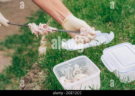 Männliche Hände in weißen Handschuhen, die rohes Fleisch in Marinade auf Spieße zum Kochen von Grills festziehen Stockfoto