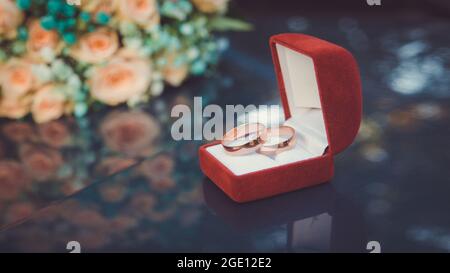 Hochzeitsringe aus Gold auf dem Hintergrund eines Bouquets von Pfirsichrosen in einer roten Box aus der Nähe Stockfoto