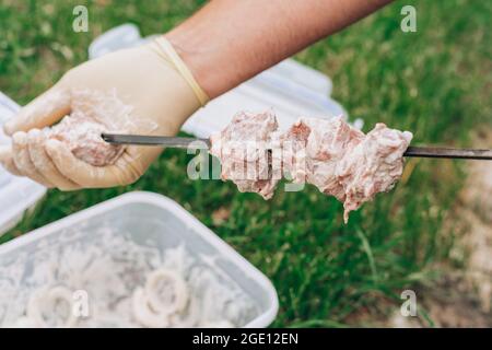 Männliche Hände in weißen Handschuhen, die rohes Fleisch in Marinade auf Spieße zum Kochen von Grills festziehen Stockfoto