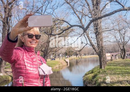 Erwachsene Frau mit Sonnenbrille, die an einem sonnigen Tag ein Selfie im Park mit einer Lagune hinter sich nimmt. Stockfoto