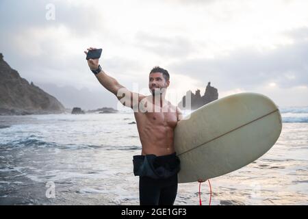 Sportlicher junger Surfer macht ein Selfie am Strand, sportlicher Typ hält ein Surfbrett und steht während des Sonnenuntergangs an einem tropischen Strand - sportlich tausendjährig Stockfoto