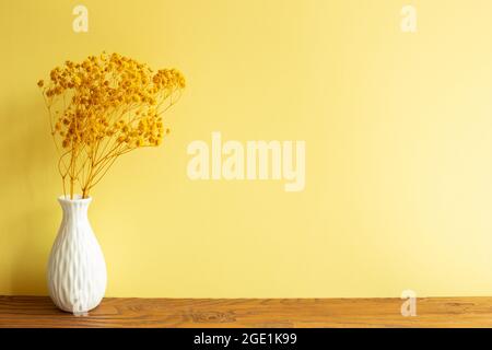 Bund von gelben Baby Atem Blumen in weißer Vase auf Holztisch. Gelbe Wand Hintergrund. Home Interieur Stockfoto