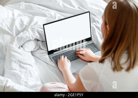 Junge Frau im Schlafanzug sitzt auf dem Bett mit Laptop mit leerem Modell Vorlage auf dem Bildschirm. Remote-Arbeit vom heimischen Arbeitsplatz im Bett mit einem Laptop Stockfoto