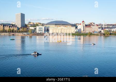 Am Frühlingsabend segeln zahlreiche Katamarane und Boote entlang des ruhigen Flusses gegen moderne Gebäude am Ufer Stockfoto