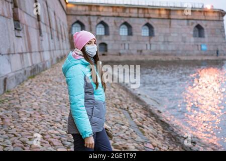 Eine junge Frau kaukasischer Ethnie geht in warmer Oberbekleidung und einer Schutzmaske durch die Stadt am Fluss, im Herbst geht sie während einer Pandemie um die Stadt Stockfoto