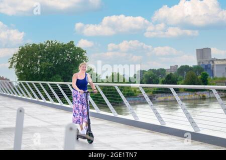 Eine blonde Frau mittleren Alters, die im Sommer bei Sonnenschein mit Copyspace auf einem Elektroroller über eine Brücke über einen städtischen Fluss fährt Stockfoto