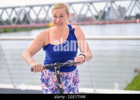 Aktive attraktive Frau mittleren Alters, die auf einer Brücke über einen Fluss mit einem Elektroroller fährt und fröhlich lächelt, während sie auf einem sonnigen Su zur Seite schaut Stockfoto