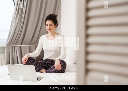 Die Frau, die mit gekreuzten Beinen vor einem Laptop auf dem Bett sitzt, während sie in einem hellen Raum auf seine Kamera blickt Stockfoto